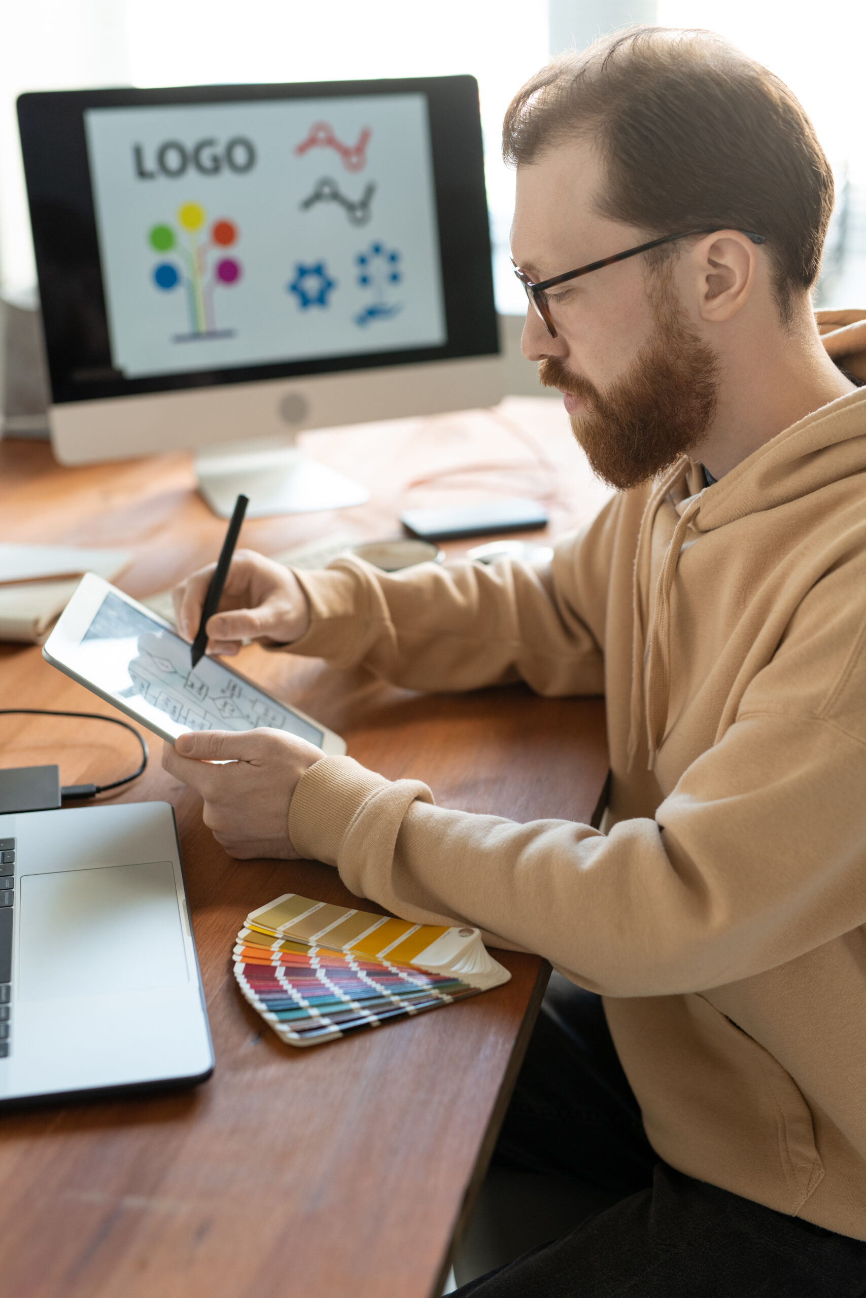 Concentrated bearded man in hoodie sitting at wooden table and using tablet while working on brand design